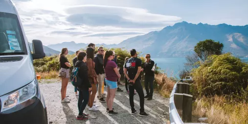Tour group gathered at a lookout with a guide talking, overlooking Lake Wakatipu and the mountains