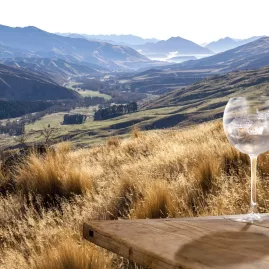 Bottle of Source Gin and glass set on a wooden table overlooking the Cardrona Valley and surrounding mountains.
