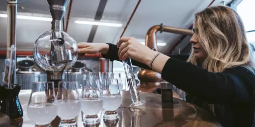 Woman measuring distillate into glasses inside Cardrona Distillery, surrounded by copper stills.