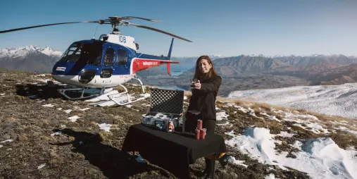 Woman presenting a gin cocktail next to a helicopter on a snowy mountaintop in the Cardrona Range.