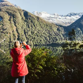 Guest in red jacket taking photos of lush green hills and snow-capped mountains on the Milford road