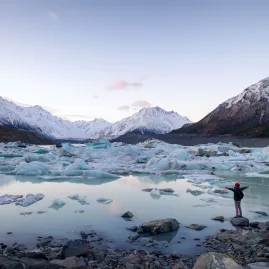 Person standing at the edge of Tasman Glacier Lake surrounded by floating icebergs.
