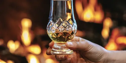 Hand holding whisky glass in front of warm fireplace