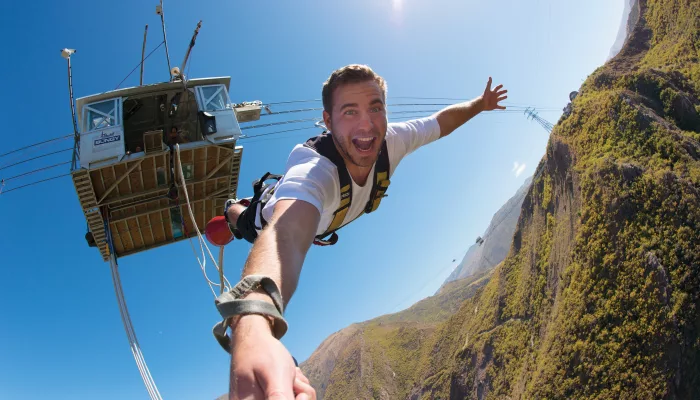 A smiling man captured mid-fall during a bungy jump from the Nevis Bungy platform high above a rugged Central Otago valley.