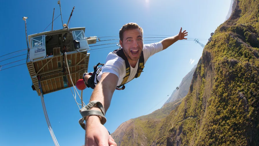 A smiling man captured mid-fall during a bungy jump from the Nevis Bungy platform high above a rugged Central Otago valley.