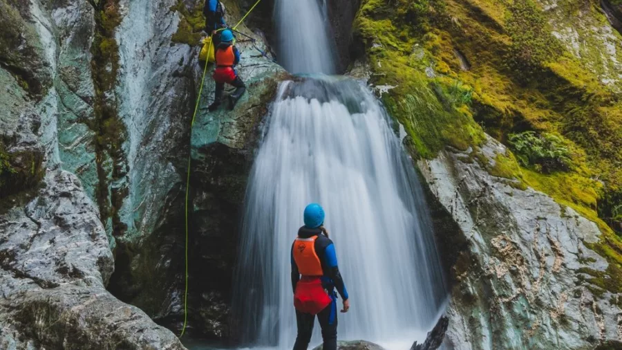 A canyoner prepares to abseil down a tall, cascading waterfall surrounded by mossy rock during the Routeburn experience.