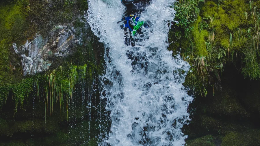 A participant slides feet-first down a waterfall into a canyon pool as a guide watches from above in the Routeburn area.
