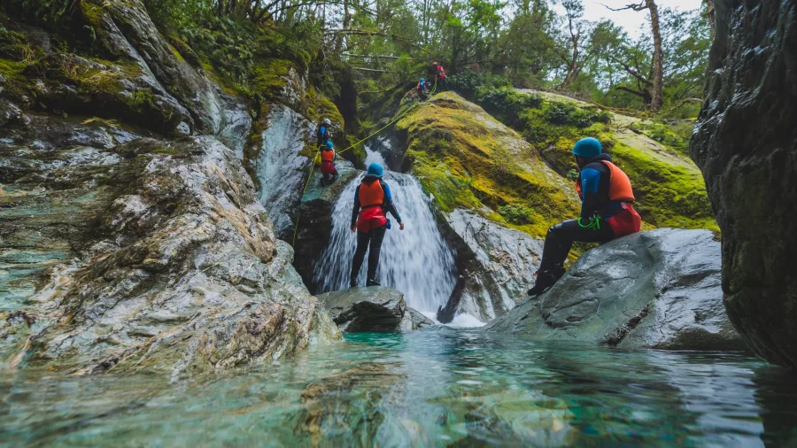 A group of canyoners descend a small waterfall in the Routeburn canyon, with one person in mid-descent and others watching from the rock pools below.
