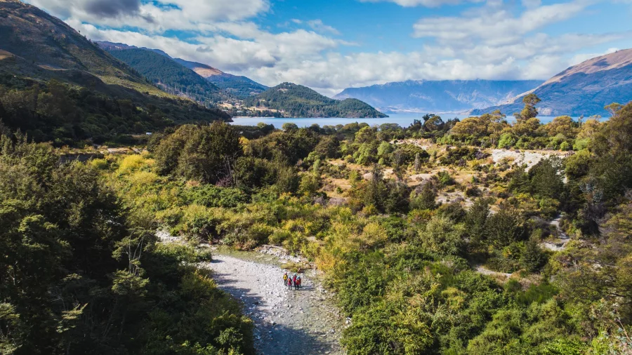 A small group of canyoners walks along a streambed surrounded by lush bush and views of Lake Wakatipu in Queenstown.