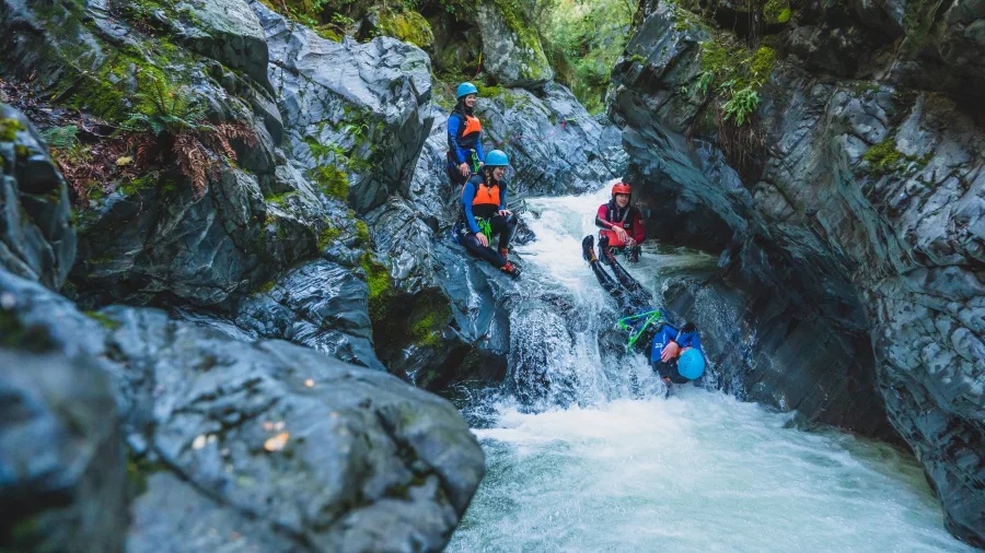 A canyoning participant prepares to slide head first into a rapid stream while a guide offers direction in Queenstown’s canyon.