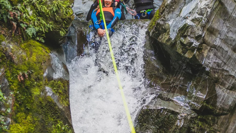 Two participants and a guide prepare to descend down a narrow waterfall using a rope in a Queenstown canyon.