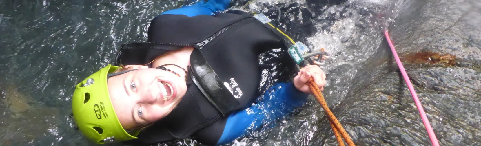 A woman in canyoning gear lies back in a water pool, smiling at the camera after a descent.