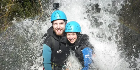 A smiling couple poses together under a waterfall during a canyoning adventure in Gibbston Valley, wearing wetsuits and helmets.