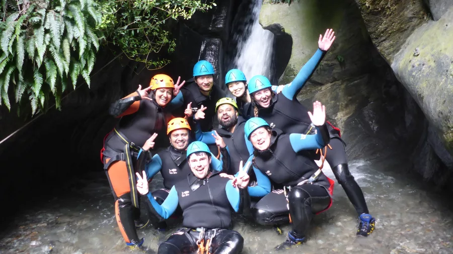 A cheerful group of canyoning participants pose for a photo at the base of a waterfall, flashing peace signs in wetsuits and helmets.