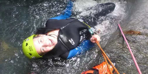 A woman in canyoning gear lies back in a water pool, smiling at the camera after a descent.