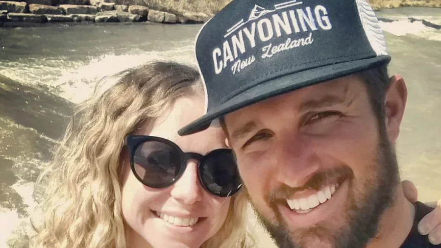 A smiling couple wearing matching Canyoning New Zealand branded t-shirts and hat pose for a selfie by the water.