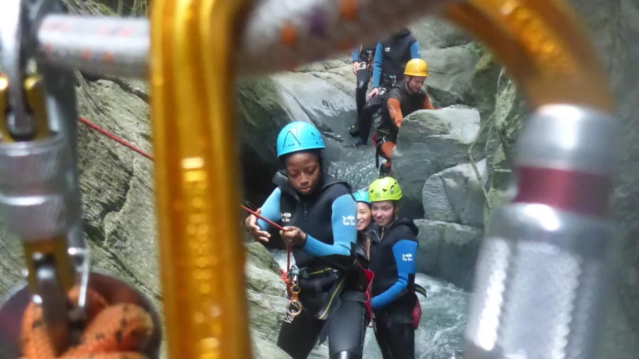 A close-up of safety karabiners in focus, with a group of canyoners blurred in the background, waiting their turn.
