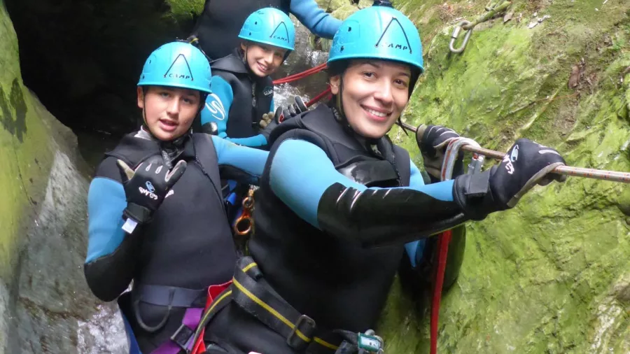 A group of canyoners navigates through a mossy, narrow canyon path with ropes and helmets.
