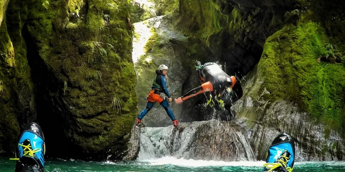 A canyoning participant leaps into a pool as a guide stands ready to assist, surrounded by mossy canyon walls.