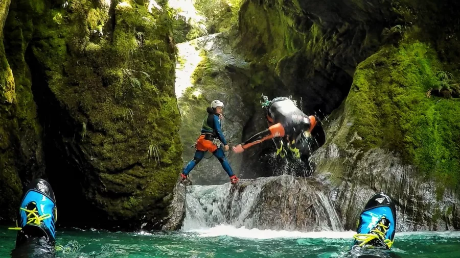 A canyoning participant leaps into a pool as a guide stands ready to assist, surrounded by mossy canyon walls.