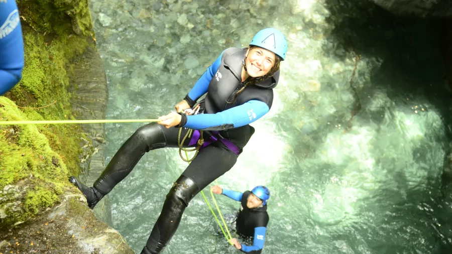 A smiling woman abseils down a mossy cliff into a crystal-clear canyon pool below.