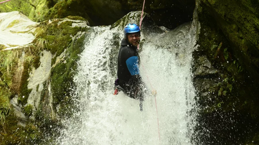 A man abseils down a powerful waterfall with a big smile, surrounded by mossy rock.