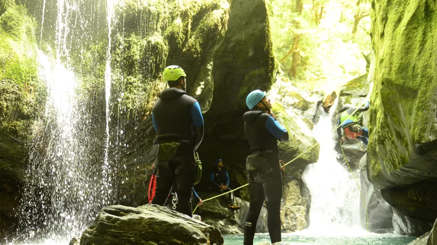 Two canyoners watch a teammate abseiling into a canyon pool beside a waterfall.