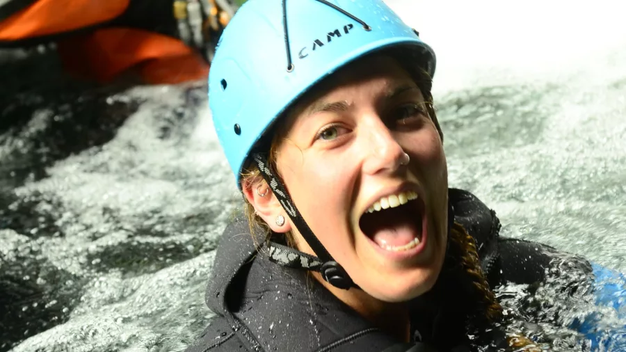 A woman in a wetsuit and helmet smiles brightly while floating in canyon water.