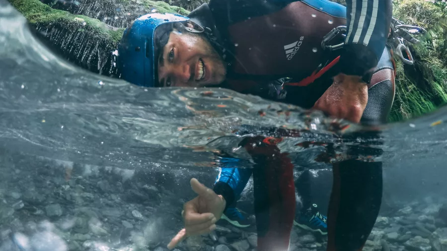 A canyoner smiles and gestures playfully while partially submerged in a clear mountain stream.
