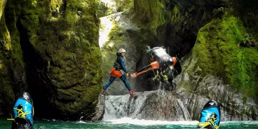 A canyoning participant leaps into a pool as a guide stands ready to assist, surrounded by mossy canyon walls.