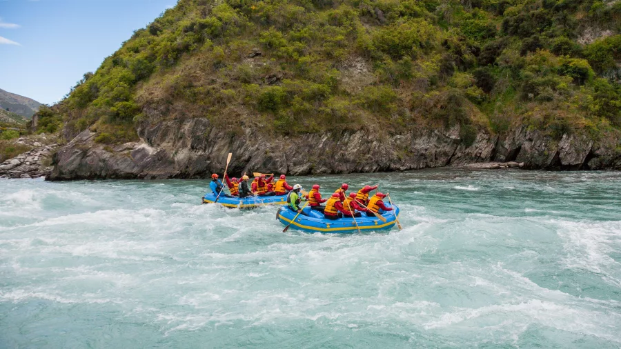 Two blue rafts float through a gentle section of the Kawarau River, framed by lush green hills.