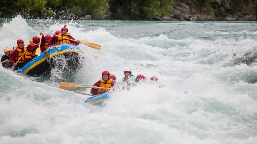 Two rafts charge into a frothy rapid side by side, with paddlers bracing and splashing through the water.