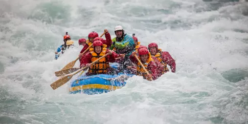 Rafters paddle hard through white water, with waves crashing over the raft’s front edge.