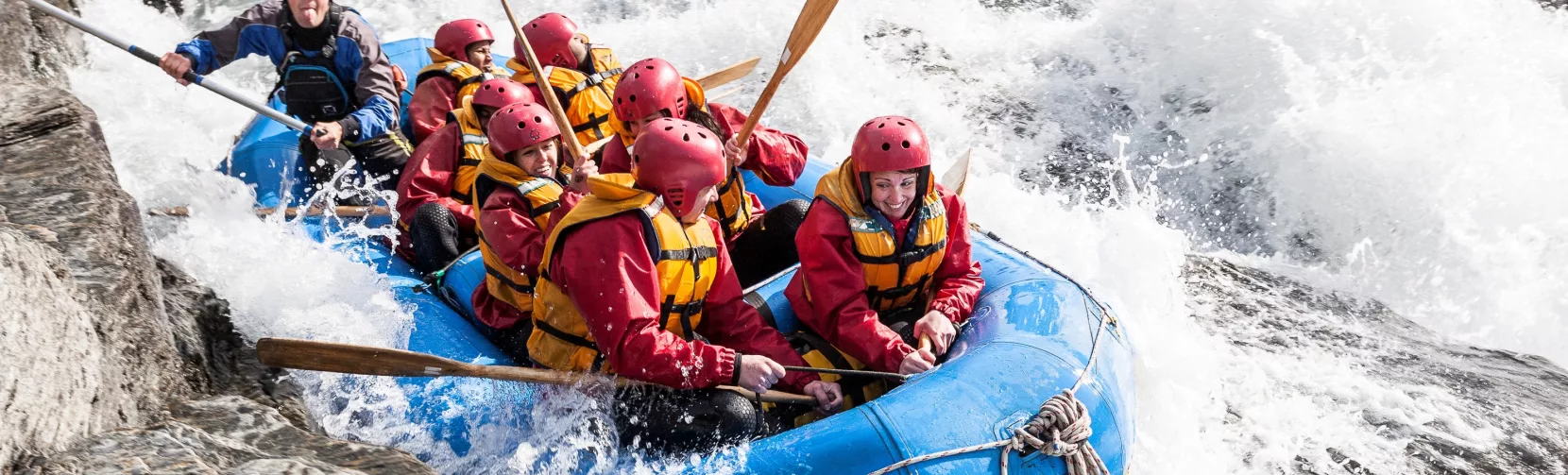 Rafters in red jackets and helmets navigate a powerful rapid beside jagged rocks on the Shotover River.
