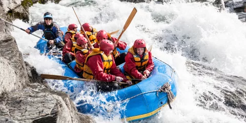 Rafters in red jackets and helmets navigate a powerful rapid beside jagged rocks on the Shotover River.