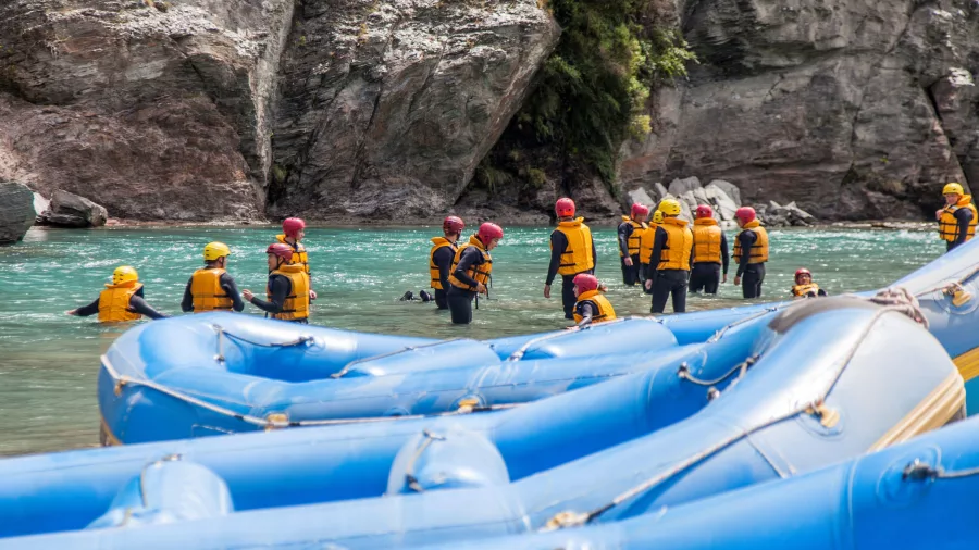 Rafters in helmets and life vests prepare for their Shotover river journey beside inflatable blue rafts.