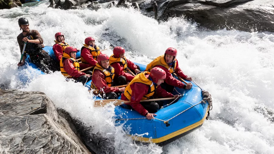 Rafters wearing red and yellow gear plunge through intense whitewater rapids in a blue raft.