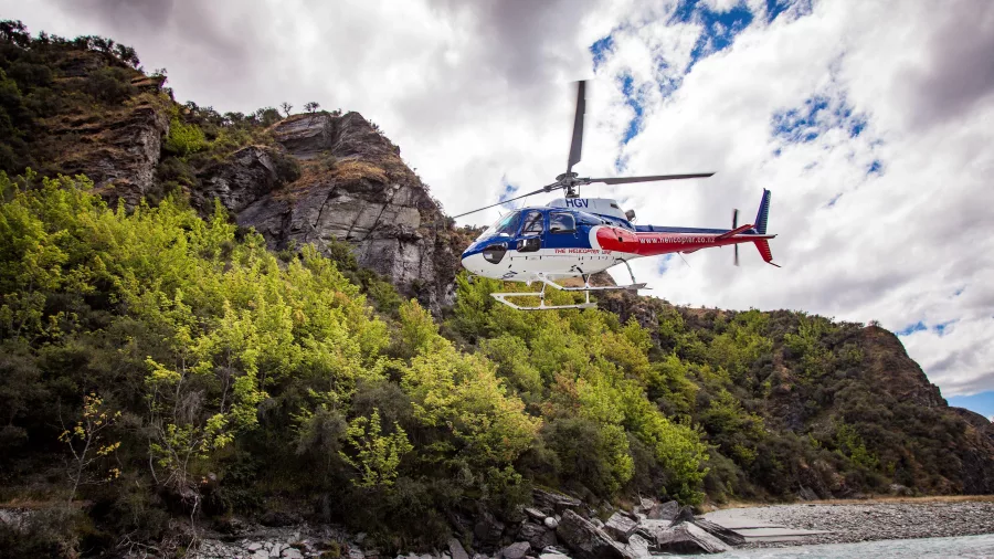 A helicopter takes off beside a riverbank surrounded by cliffs and forest, following a rafting trip.
