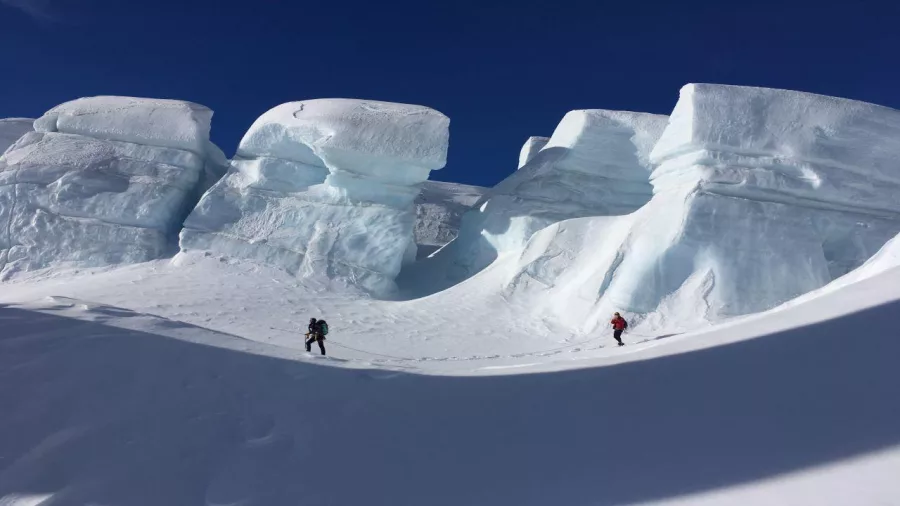 Two hikers walking through deep snow below towering ice cliffs on a Mount Cook glacier tour