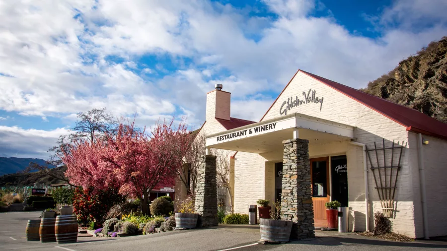 Exterior view of Gibbston Valley Restaurant & Winery with cherry blossoms, wine barrels, and mountain surrounds.