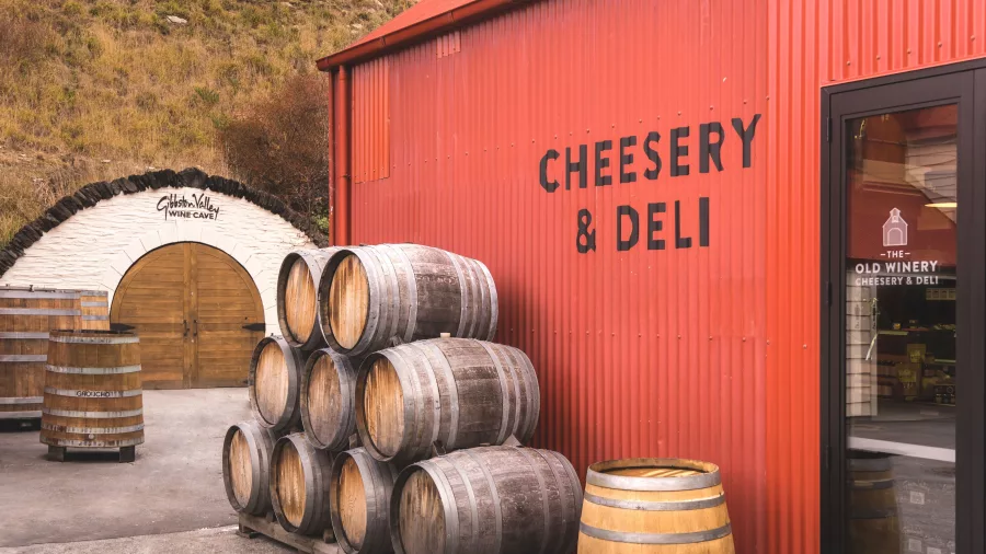 Exterior of Gibbston Valley Cheesery & Deli with stacked wine barrels and signage on a red building.