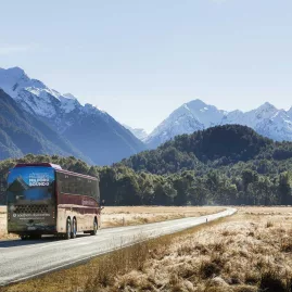 Coach driving towards Milford Sound with snow-capped mountains in the distance.