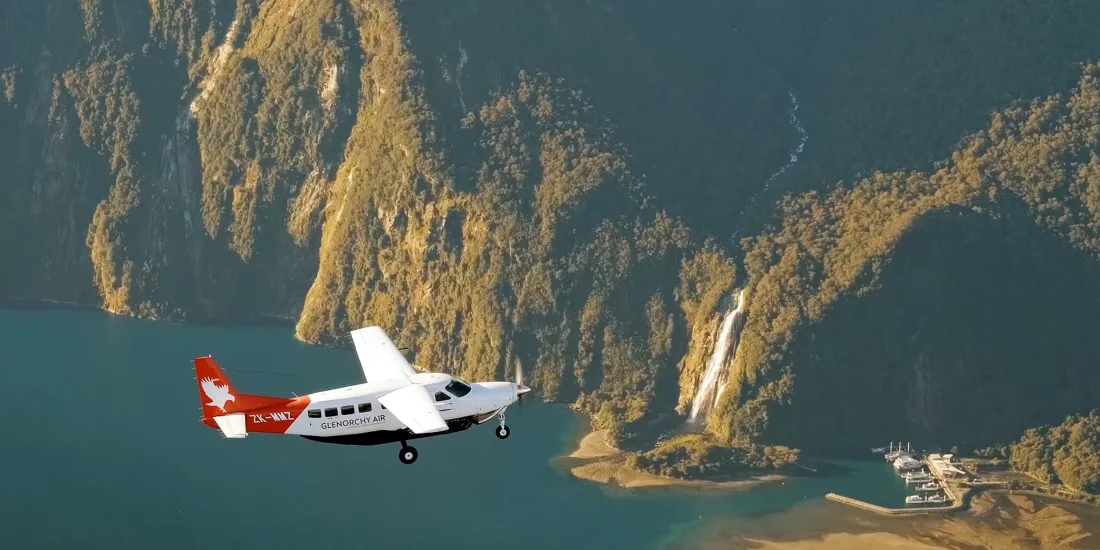 Light aircraft flying above Milford Sound with the marina, airstrip and Stirling Falls visible below in Fiordland National Park