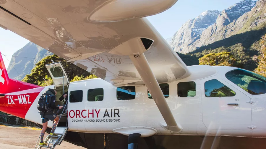Passenger boarding a Glenorchy Air flight at Milford Sound airstrip with scenic peaks in the background.