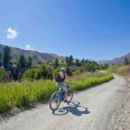 Cyclist rides a sunny gravel trail beside the river in Gibbston Valley