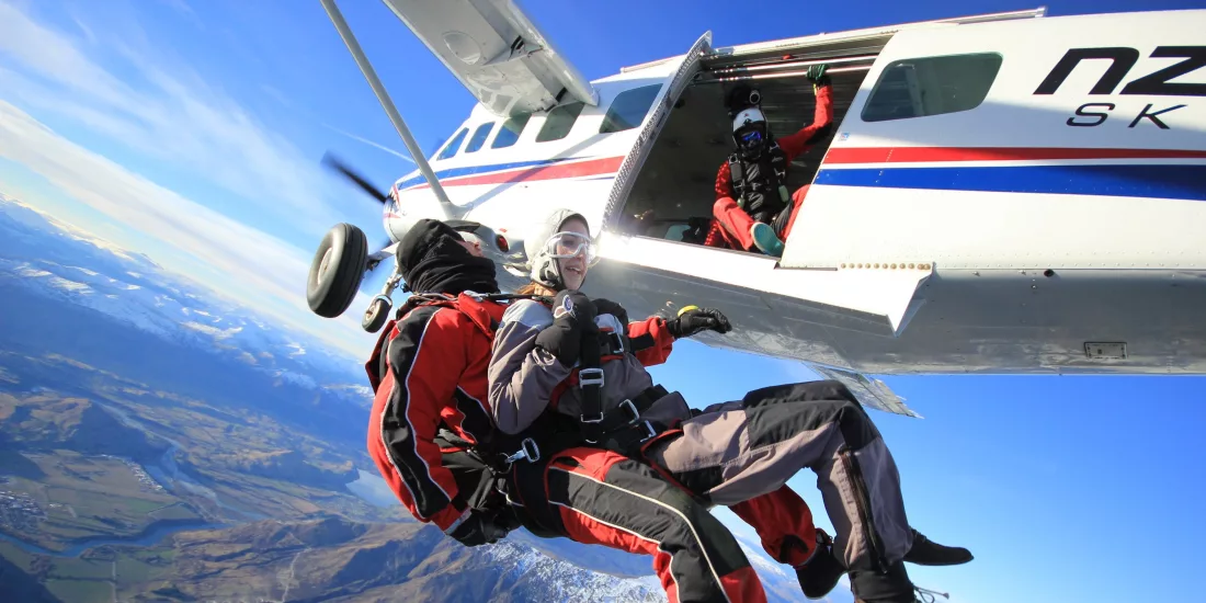 Tandem skydivers exiting a plane high above Queenstown, with mountain and lake views below.