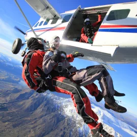 Tandem skydivers exiting a plane high above Queenstown, with mountain and lake views below.