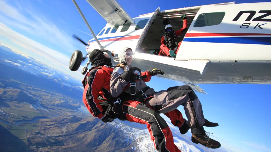 Tandem skydivers exiting a plane high above Queenstown, with mountain and lake views below.