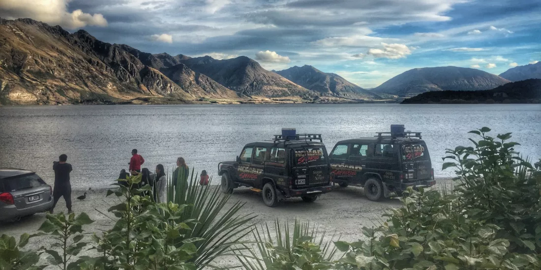 Nomad Safaris Land Rovers parked beside Lake Wakatipu with scenic mountains in the background