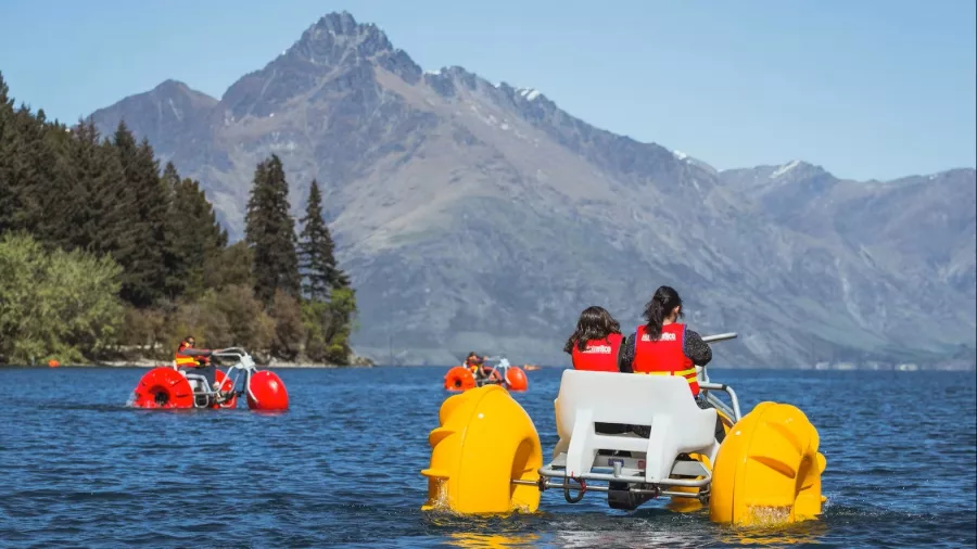 People riding red and yellow aqua bikes on Lake Whakatipu with mountain backdrop